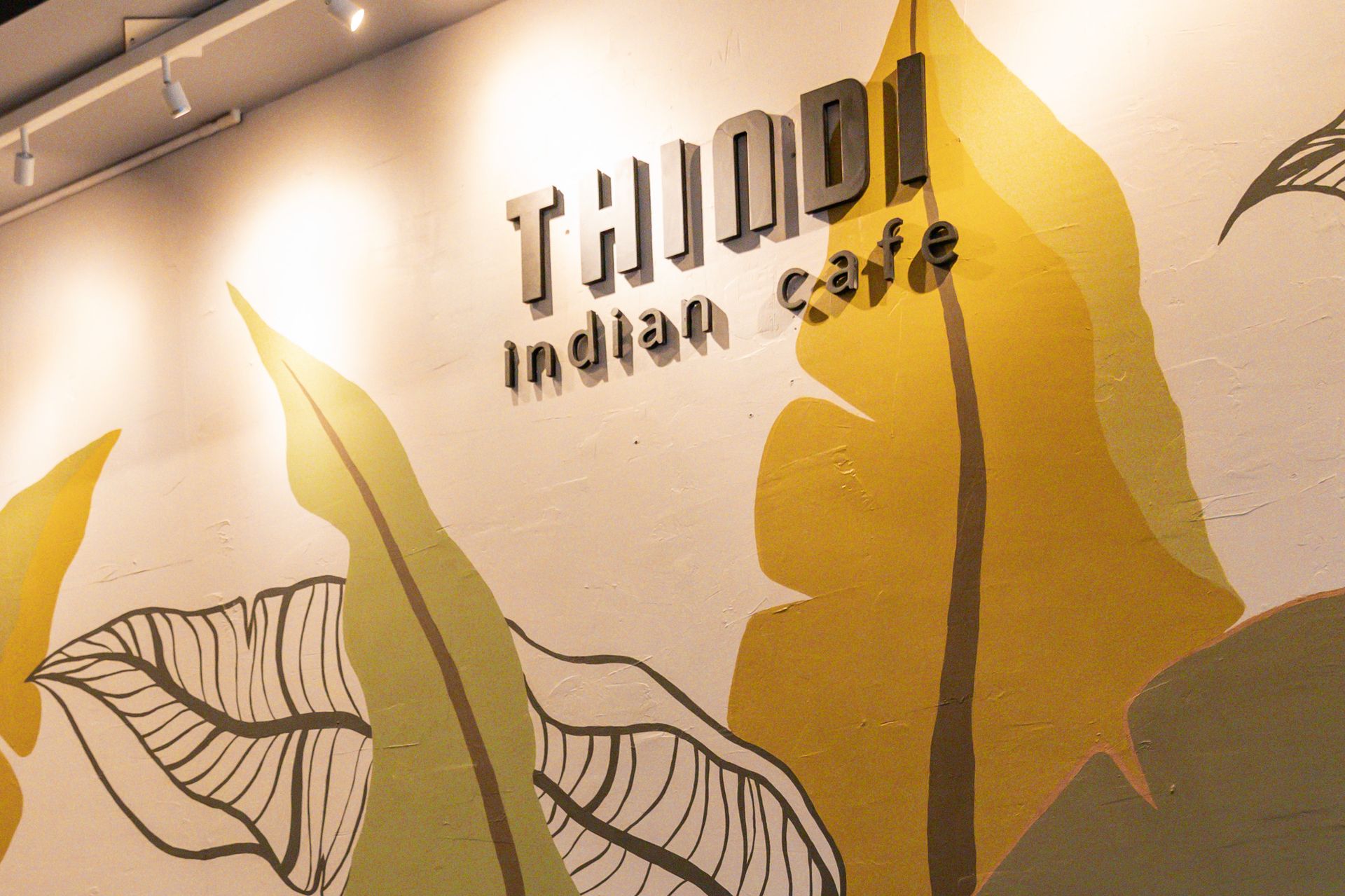 The painted leaf mural inside Thindi Cafe on College Street, with the cafe's sign above a table and two chairs
